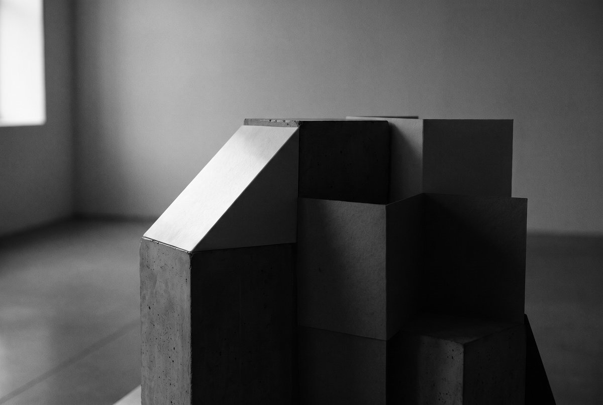 Editorial illustration — an unfinished sculptural structure held in shadow, with one partially illuminated edge. Conceptual monochrome study of patient construction and the pressure of an unfinished idea. Not a documented project or person.