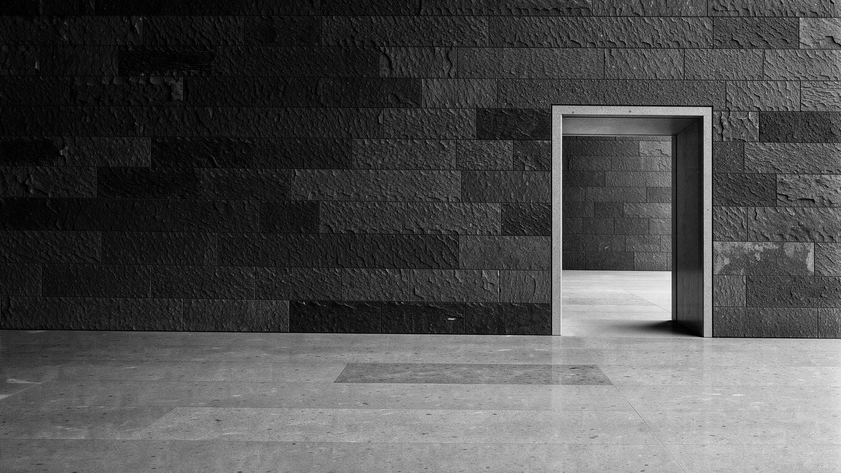 Editorial illustration — a clean rectangular doorway cut through a thick dark stone wall, with soft daylight on the other side. Conceptual monochrome study of a credible route out — the ability to leave. Not documentary.