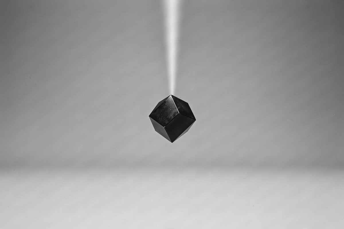 Editorial illustration — a single small polished geometric form suspended in the exact centre of a vast empty studio field; one thin beam of light converging on it. Conceptual monochrome study of the valuable unit of knowledge within an open field. Not documentary.